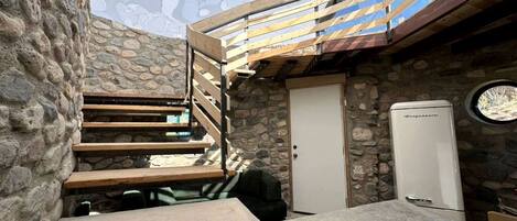 Una habitación con paredes de piedra, una escalera de madera, una puerta y un electrodoméstico blanco.