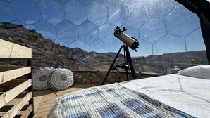 Ein Teleskop auf einem Stativ in einem Raum mit Holzboden, einem Bett und Blick auf eine Berglandschaft durch ein sechseckiges Fenster.