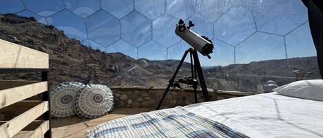A telescope on a tripod in a room with a wooden floor, a bed, and a view of a mountainous landscape through a hexagonal window.