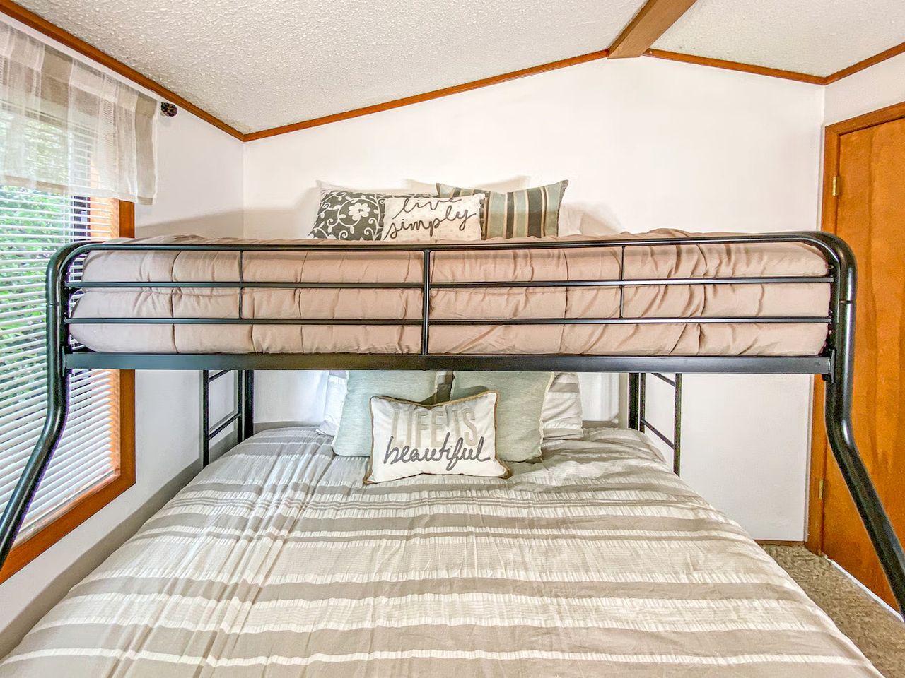 A bunk bed with two levels, a window with blinds, and a wall with a decorative pillow.