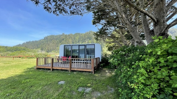 A modern cabin with a deck, surrounded by lush greenery and a forested backdrop.