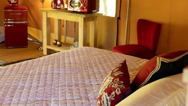 A tented room with a bed, a table with a radio, a chair, and a lamp.