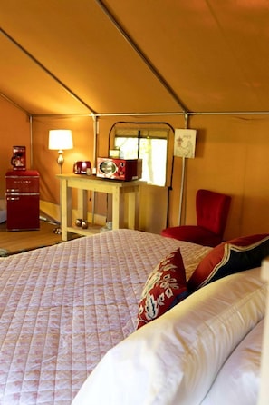 A tented room with a bed, a table with a radio, a chair, and a lamp.