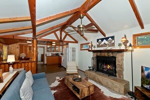 A spacious living room with a fireplace, a sofa, a coffee table, and a dining area with wooden cabinets and a kitchen in the background. - Forest Cottage with Outdoor Jacuzzi and Picnic Space in California (Big Bear Lake)