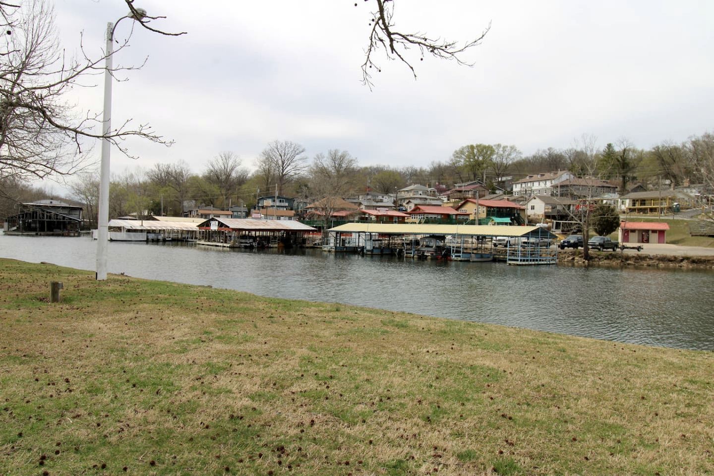 A riverside view with docked boats, a grassy bank, and residential buildings in the background.