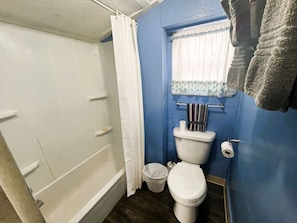 A bathroom with a white bathtub, a toilet, a small trash bin, and a window with a curtain. - One Bedroom Lake Cabin on Lake Taneycomo, Rockaway Beach, Missouri (Rockaway Beach)