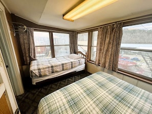 A small room with two beds, a window with curtains, a radiator, and a view of a parking area.