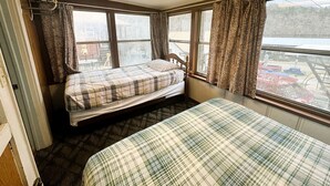 Ein kleines Zimmer mit zwei Betten, einem Fenster mit Vorhängen, einem Heizkörper und Blick auf einen Parkplatz.