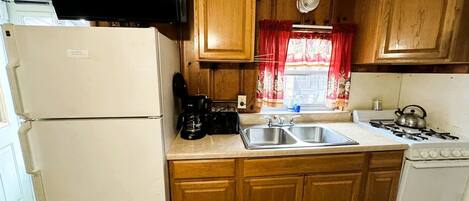 A compact kitchen with wooden cabinets, a white refrigerator, a stove, a sink, and a television mounted above the counter.
