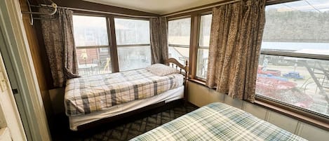 A small room with two beds, a window with curtains, a radiator, and a view of a parking area.