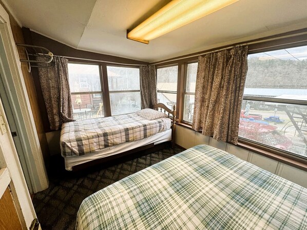 A small room with two beds, a window with curtains, a radiator, and a view of a parking area.