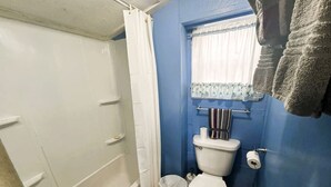Ein Badezimmer mit einer weißen Badewanne, einer Toilette, einem kleinen Mülleimer und einem Fenster mit Vorhang.