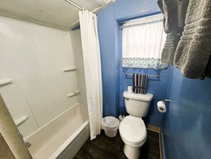 Un baño con una bañera blanca, un inodoro, un cubo de basura pequeño y una ventana con cortina.