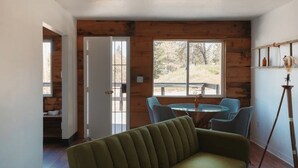 Una habitación con un sofá verde, una mesa redonda con sillas, suelo de madera y un amplio ventanal con vistas a árboles.