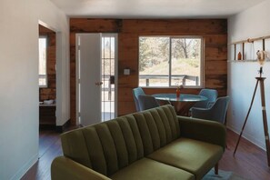 Ein Zimmer mit einem grünen Sofa, einem runden Tisch mit Stühlen, Holzboden und einem großen Fenster mit Blick auf Bäume.