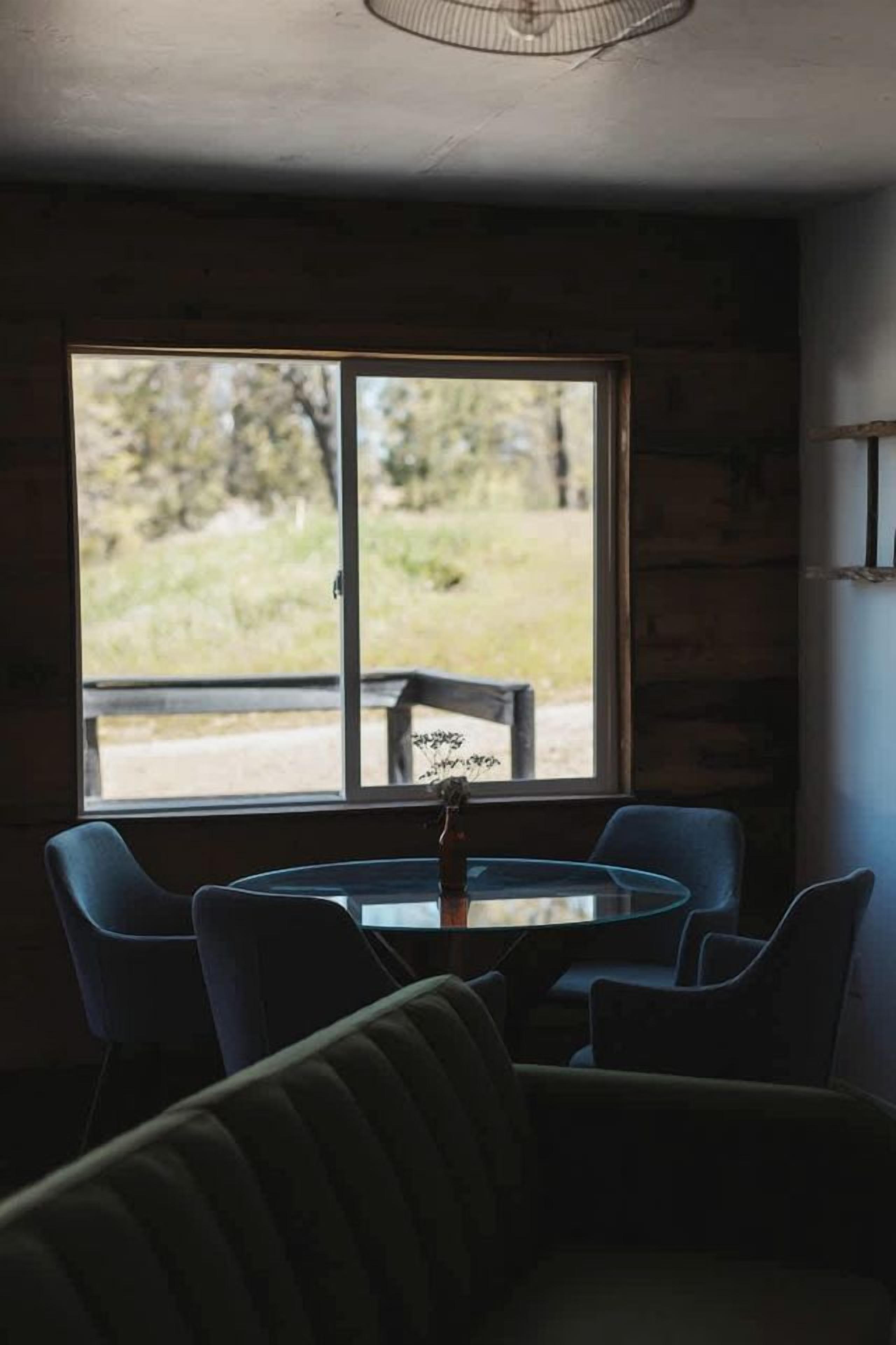 Quarto com mesa redonda e cadeiras, janela com vista para área verde e ventilador de teto.