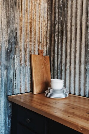 Una mesa de madera con platos blancos apilados y una tabla de cortar de madera frente a una pared de metal corrugado.