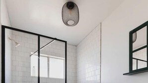 Un baño con cabina de ducha de vidrio, paredes cubiertas de azulejos blancos, un lavabo blanco y un espejo sobre él.