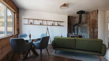 A cozy room with a wooden floor, a green sofa, a dining table with chairs, a standing lamp, and a rustic fireplace.