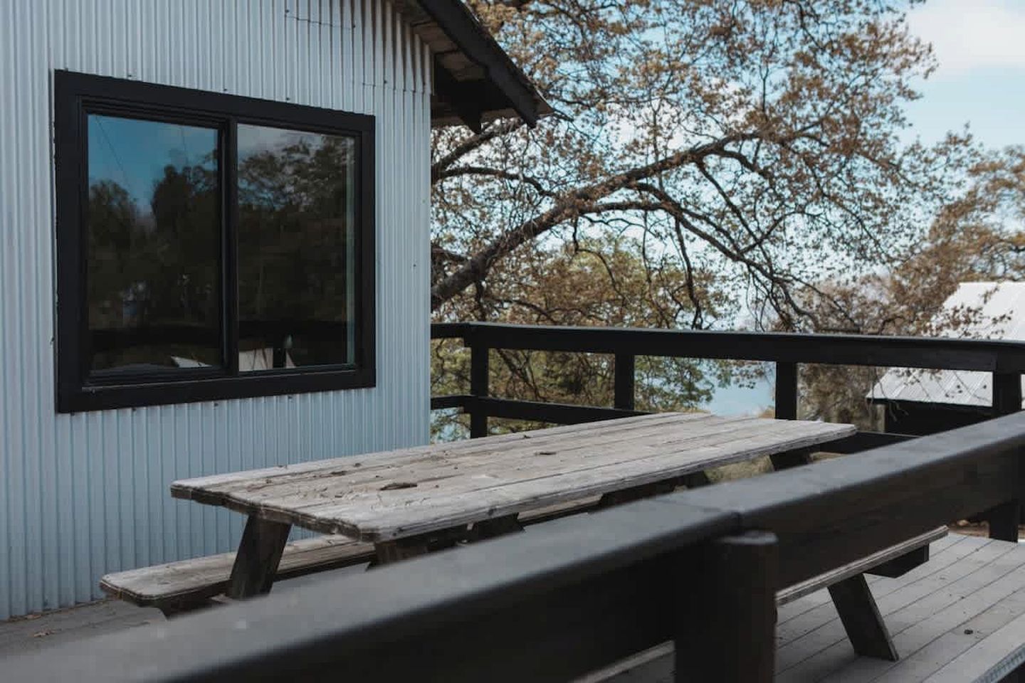 Uma mesa de piquenique de madeira numa varanda com guarda-corpo de metal, uma janela e um edifício com revestimento de metal ondulado.