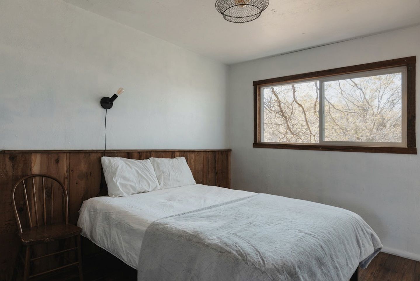 A bedroom with a wooden headboard, a single bed, a chair, and a window with a view of trees.