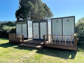 Portable toilets with a wooden deck and railing, set in a grassy area with trees in the background.