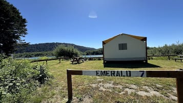 A small cabin with a white exterior and a brown roof, labeled 'EMERALDI 7', situated in a grassy area with a picnic table nearby, under a clear blue sky.