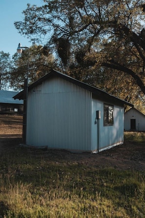A small, single-story metal building with a gabled roof, a door, and a window, surrounded by grass and trees.