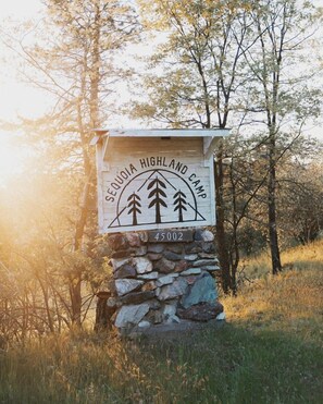 A sign for Sequoya Highland Camp with a stone base and a wooden top, surrounded by trees and grass.