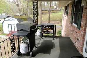 Eine überdachte Veranda mit Grill und Propangasflaschen, eine Backsteinmauer und ein kleines Gartenhaus im Hintergrund.