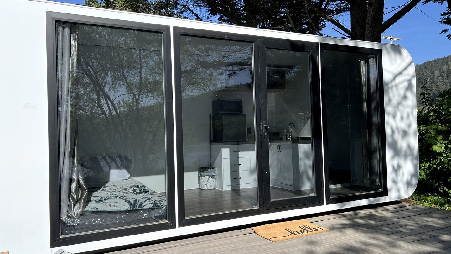 A modern, compact living space with large sliding glass doors, a visible kitchen area, and a small outdoor deck with a welcome mat.
