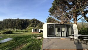 Uma motorhome moderna com varanda e cozinha, situada em um campo gramado com uma colina arborizada ao fundo.