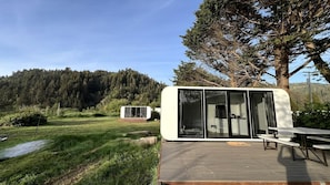 A modern RV with a deck and a kitchen area, set in a grassy field with a forested hill in the background.