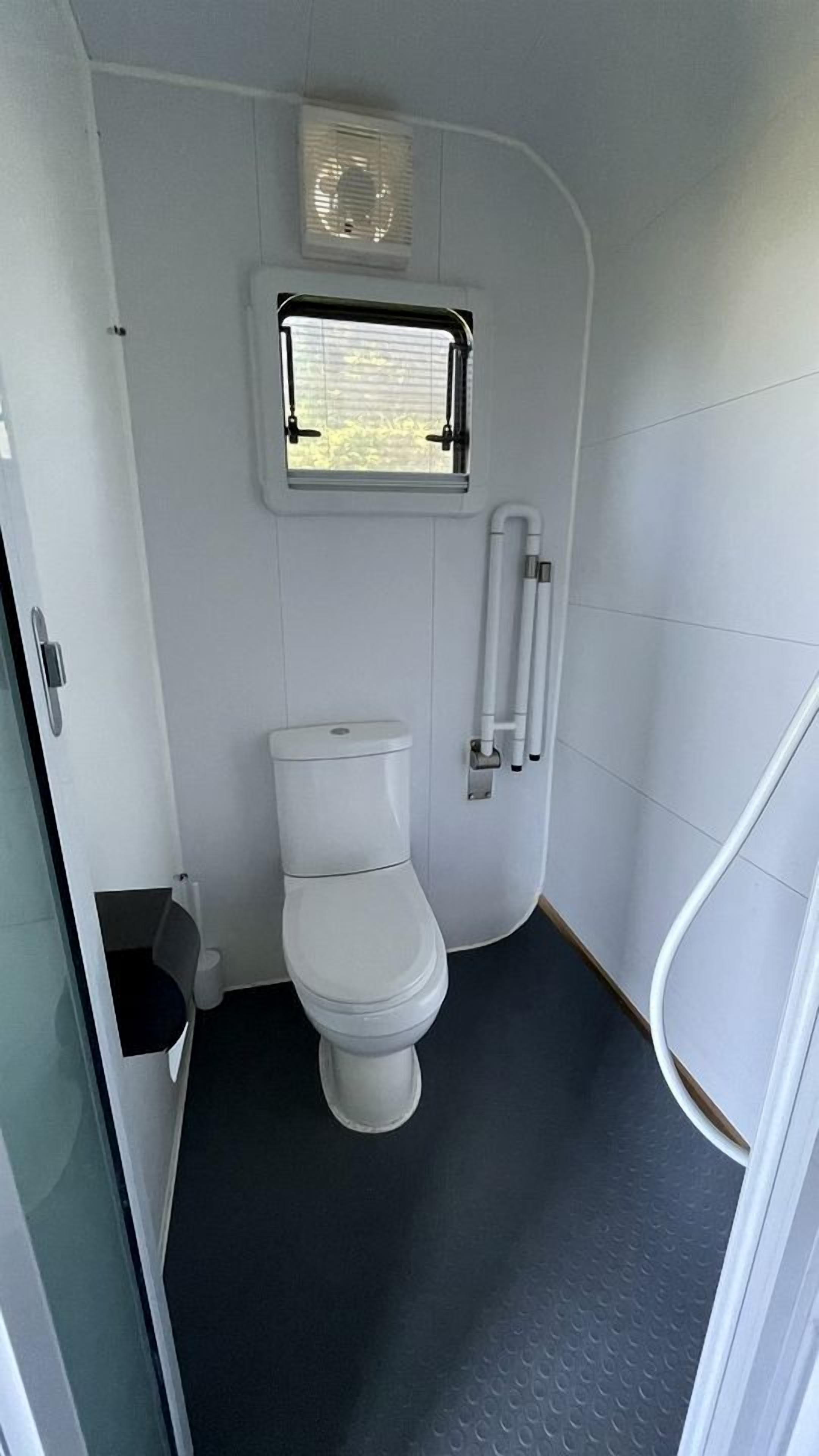 A compact bathroom with a toilet, a window, and a handrail.