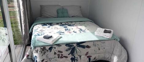 Un lit simple avec une couvre-lit à motifs floraux, une fenêtre avec des stores et une petite étagère fixée au mur.