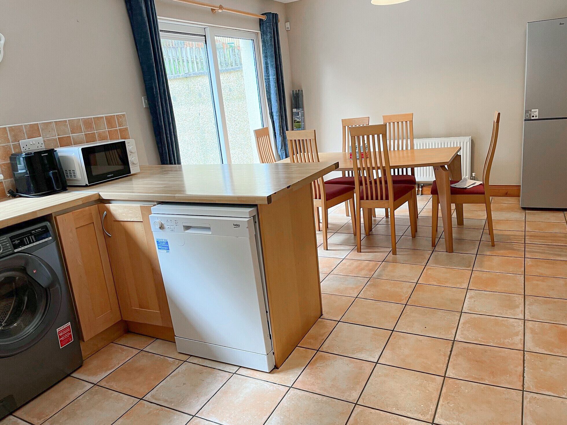 A compact kitchen with a dishwasher, microwave, and washing machine. There is a dining area with wooden chairs and a table.