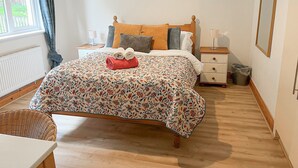 Une chambre à coucher bien rangée, avec une couvre-lit à motifs floraux, une tête de lit en bois, une table de chevet avec une lampe et un fauteuil en osier.
