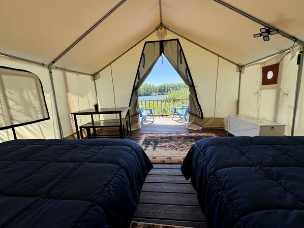 A tent with two beds, a table, and chairs, with a view of a lake and trees outside.