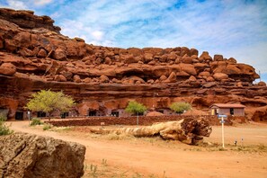 Un paysage désertique avec une grande formation rocheuse, un petit bâtiment doté de nombreuses fenêtres et une végétation clairsemée.