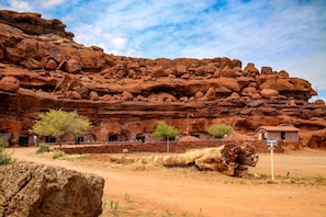 A desert landscape with a large rock formation, a small building with multiple windows, and sparse vegetation.