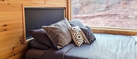 A cozy cabin interior with a bed, pillows, a small table, and a window with a view of a canyon.