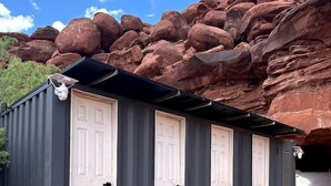 A row of small, single-room cabins with white doors, set against a backdrop of red rock formations.