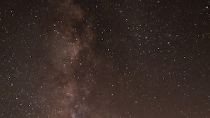 A starry night sky with the Milky Way visible, a large rock formation, and a balanced boulder.