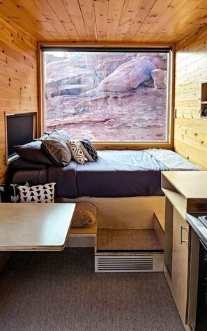 A compact cabin with a bed, a small table, and a window overlooking a rocky landscape.