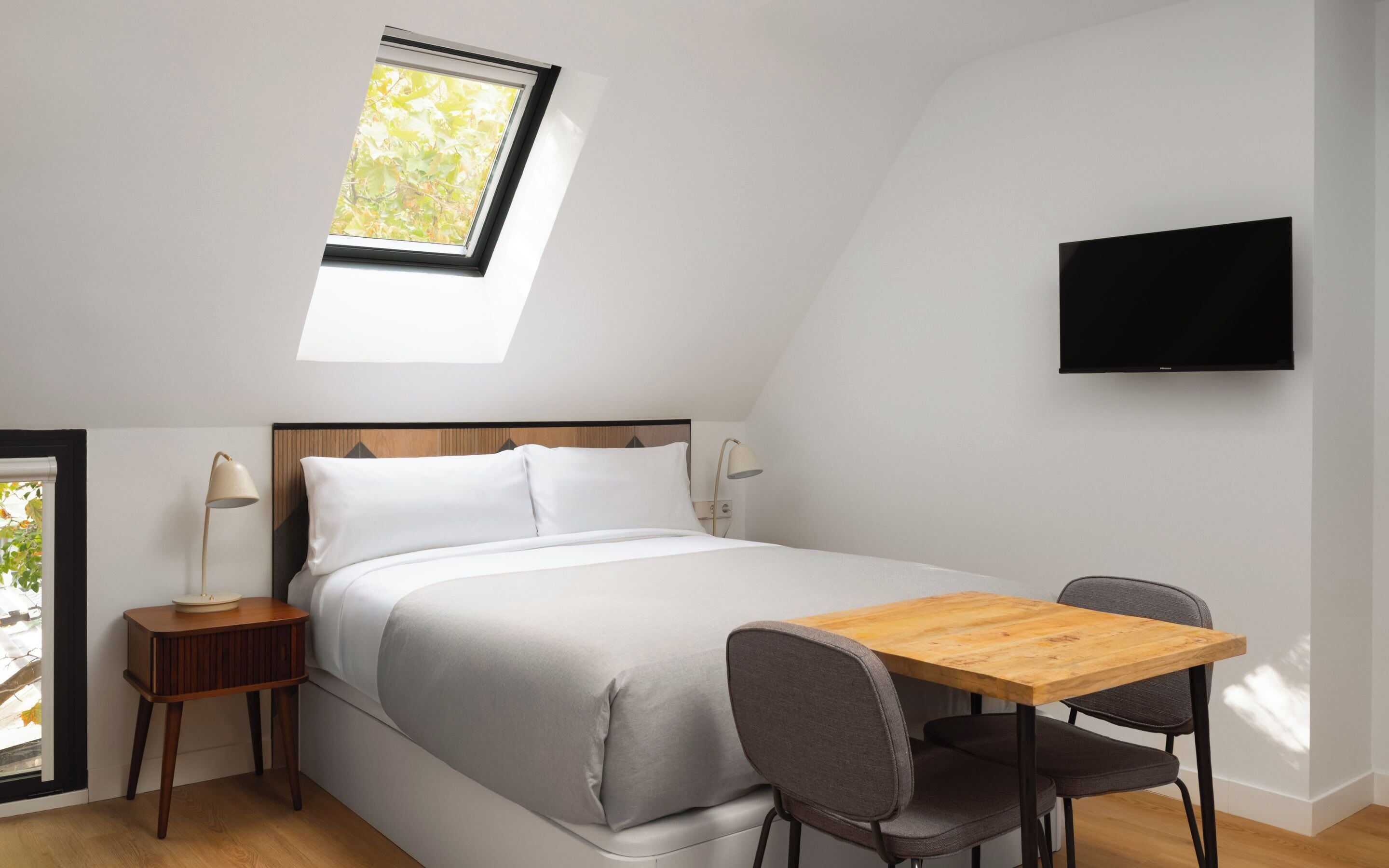 Een slaapkamer met dakraam, een bed, nachtkastje met lamp, televisie aan de muur en een klein houten tafeltje met twee stoelen.