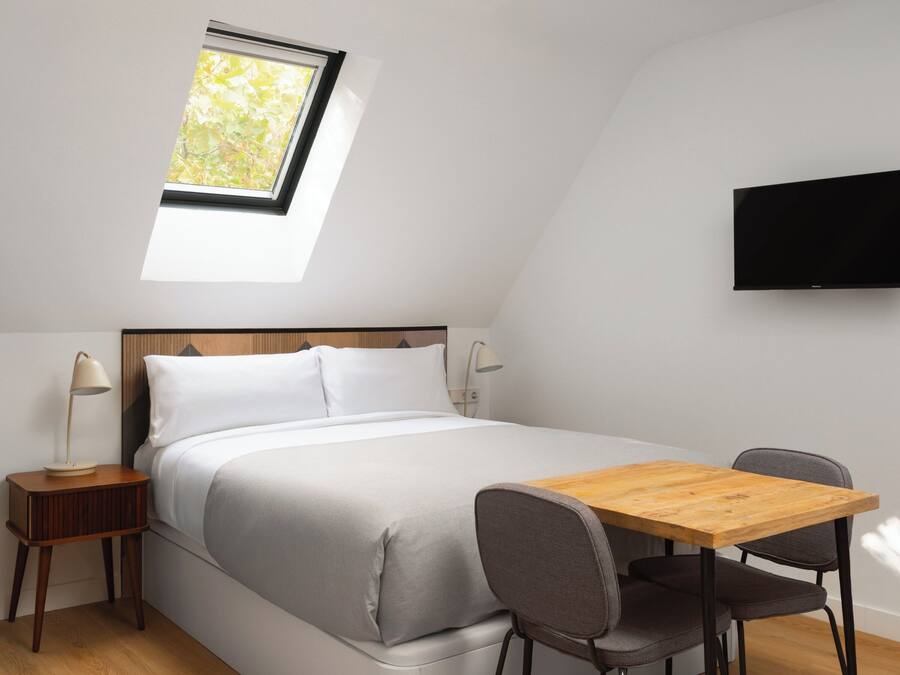 A bedroom with a skylight, a bed, a bedside table with a lamp, a TV mounted on the wall, and a small wooden table with two chairs.