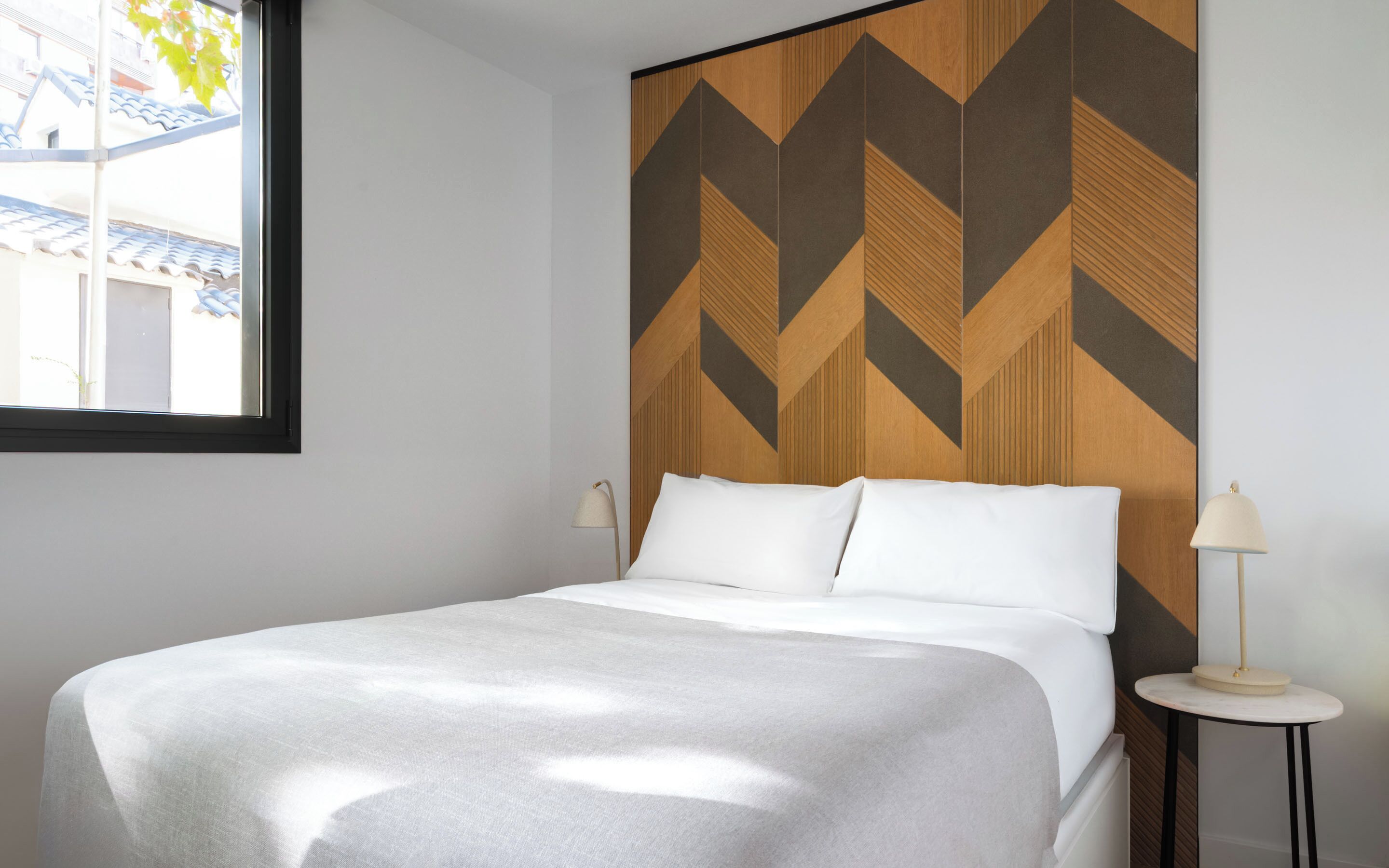 Un lit bien fait dans une chambre d’hôtel, avec une tête de lit à motifs géométriques, une lampe de chevet et une fenêtre donnant sur des immeubles.