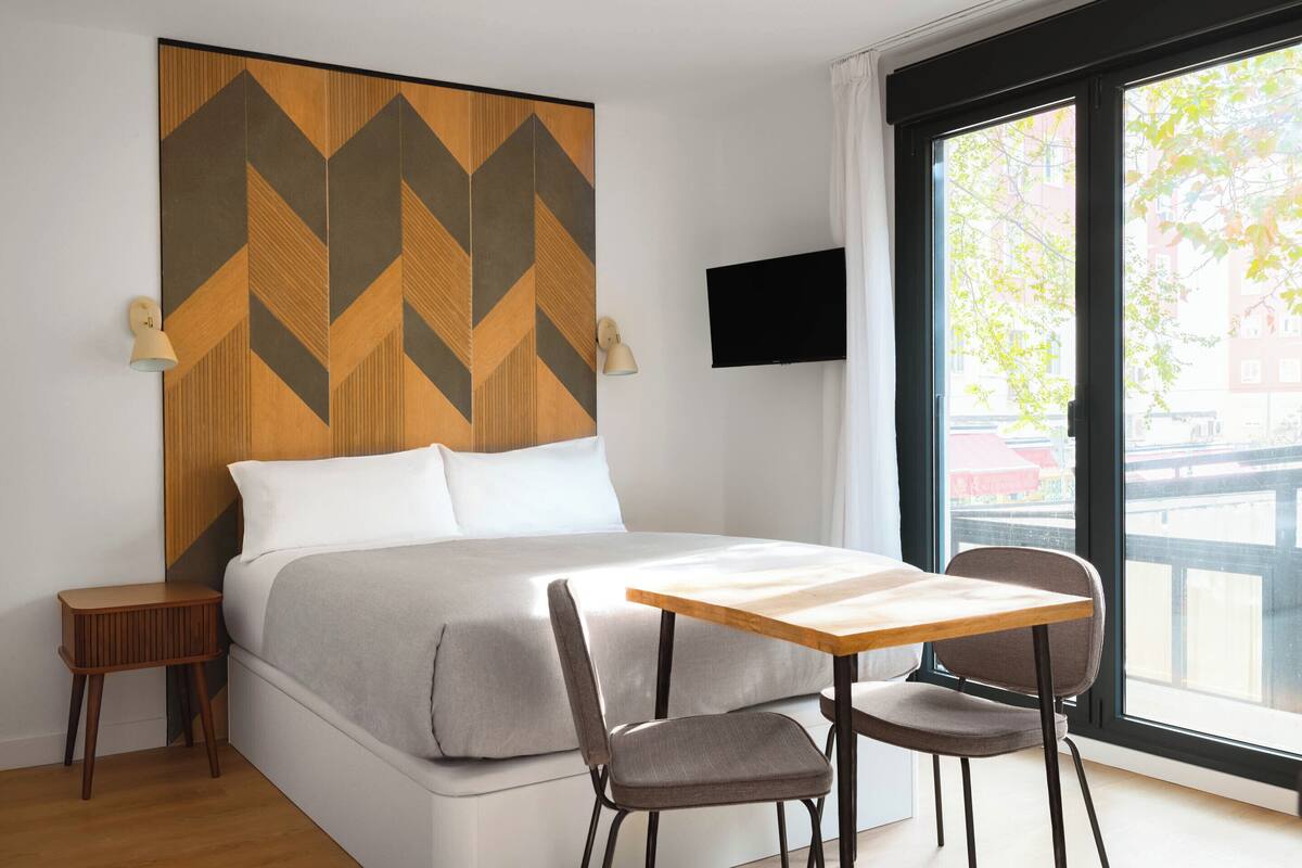 Ett modernt hotellrum med en säng, ett bord med två stolar, en TV och ett stort fönster med utsikt över byggnader och träd.