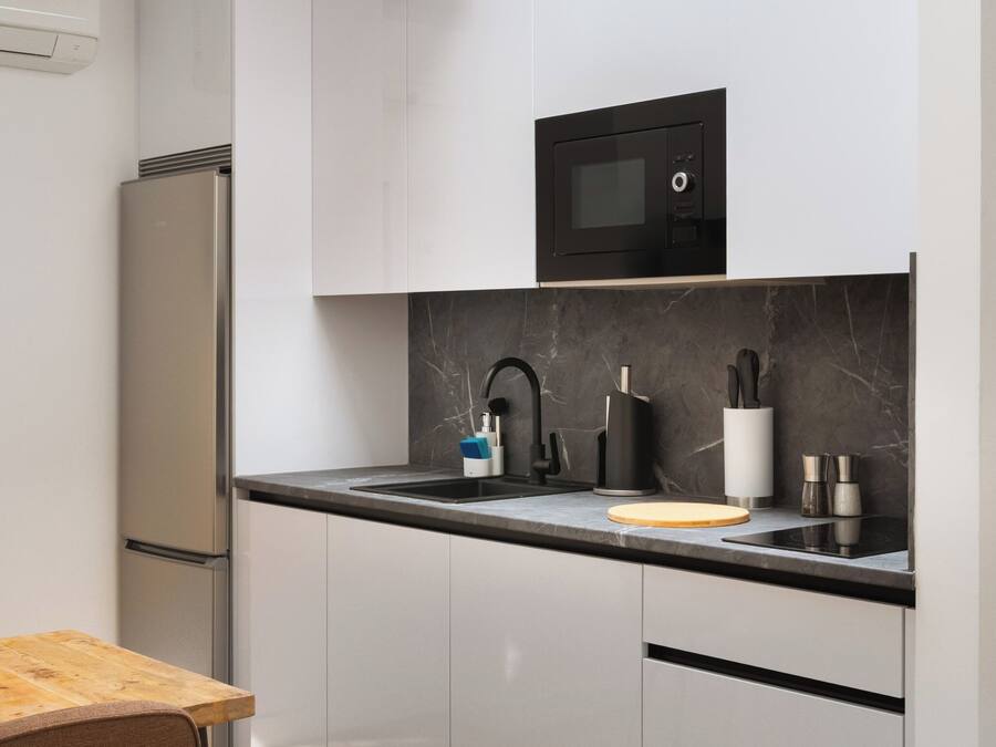 A modern kitchen with white cabinets, a black microwave, a stainless steel refrigerator, a wooden table, and a black countertop.