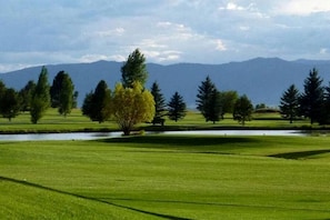 Un campo de golf con un estanque, árboles y montañas al fondo.